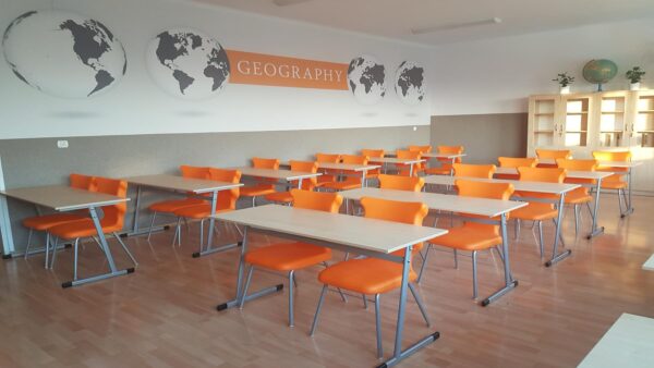Klasa szkolna z ławkami i pomarańczowymi krzesłami oraz napisem "GEOGRAPHY" na ścianie z mapami świata.