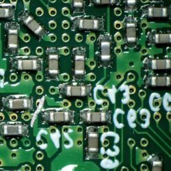 Zbliżenie na zieloną płytkę drukowaną z przylutowanymi podzespołami elektronicznymi i oznaczeniami kodowymi na ścieżkach.