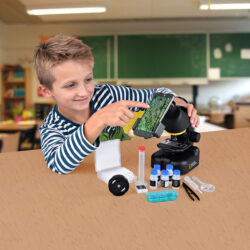 bresser-microscope-national-geographic-40-640x-03 Chłopiec ogląda próbkę pod mikroskopem z zamontowanym smartfonem, obok leżą akcesoria laboratoryjne.