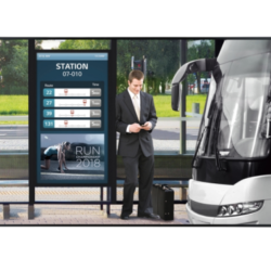 Mężczyzna w garniturze korzysta ze smartfona na przystanku autobusowym z cyfrową tablicą odjazdów.